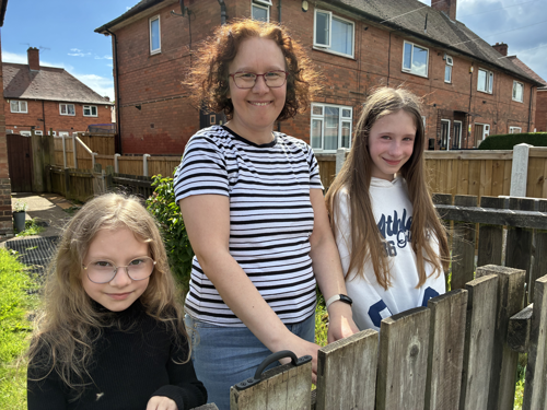 A picture of a mum and her two daughters stood at their gate smiling. Houses are in the background